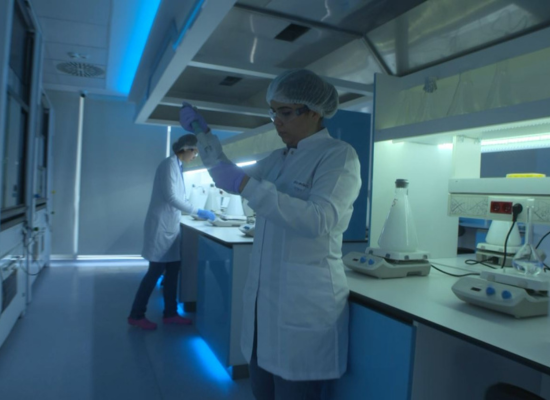 A person with a white labcoat and hairnet holding a pipette to a tube in a laboratory