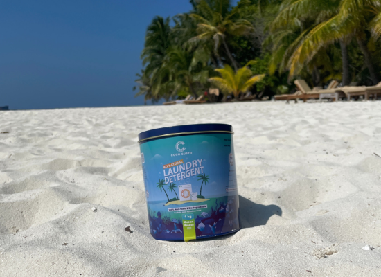 A tin of laundry detergent in the sand on a beach; palm trees and blue sky in the backgroung